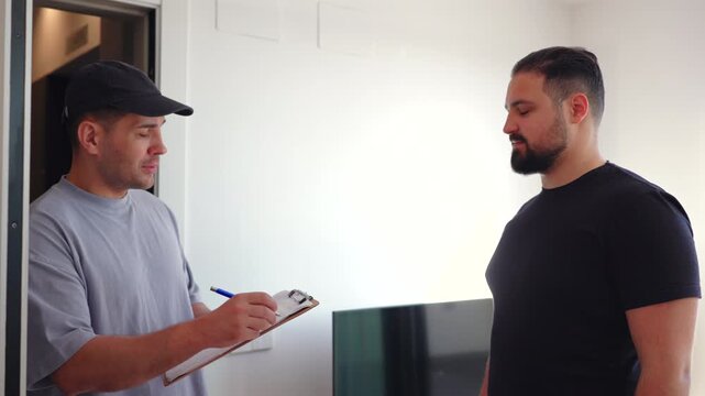 Service worker holding clipboard speaks with male client inside apartment entrance. Indoor scene reflects home inspection, maintenance consultation and professional communication.