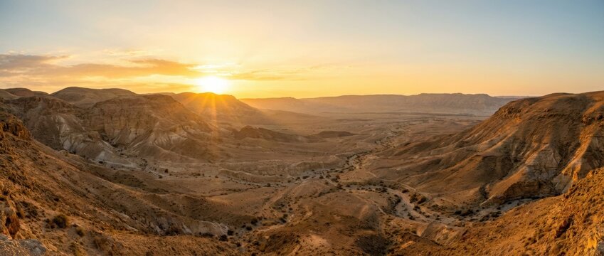 Vast arid desert landscape reveals a breathtaking panoramic view of rugged mountains and deep valleys, illuminated by the radiant golden sun setting on the horizon, creating a majestic natural scene