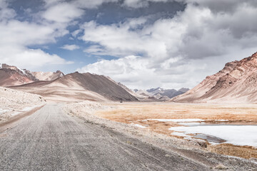Fototapeta premium Landscape in the highlands of the Tien Shan in the Pamirs of Tajikistan, rocky mountain ranges with snow and a grassy valley in cloudy weather, asphalt road, Pamir Highway