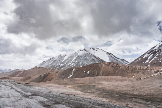 Landscape in the highlands of the Tien Shan in the Pamirs of Tajikistan, rocky mountain ranges with snow and a grassy valley in cloudy weather, asphalt road, Pamir Highway