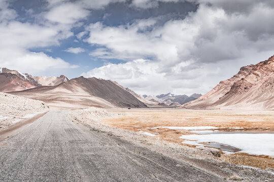 Landscape in the highlands of the Tien Shan in the Pamirs of Tajikistan, rocky mountain ranges with snow and a grassy valley in cloudy weather, asphalt road, Pamir Highway