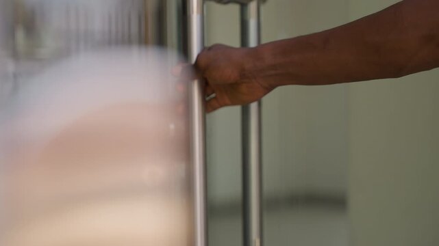 Black man pulling glass house door open with firm grip, closeup on wrist and handle interaction, tactile motion and contrast of skin and stainless metal, interior light spilling onto threshold,