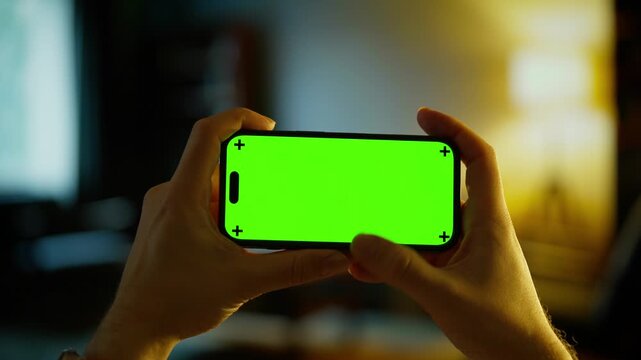 Person holding smartphone with bright green chroma key screen in dimly lit indoor environment, ready for video tracking, compositing, or customizable display content