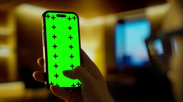 Hand holding smartphone with bright green chroma key screen in warm blurred indoor environment, showcasing mobile device display for digital design and mockup