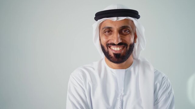 Confident young arab man with a beard wearing traditional clothing, a keffiyeh and agal, looking at the camera and smiling, expressing happiness and success on a gray studio background