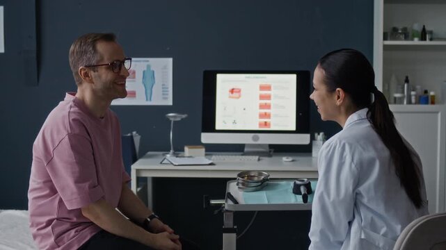 Indoor eye level shot of dermatologist consulting male patient in medical office, patient pointing at neck skin while doctor listening, computer with skin charts and examination tools visible