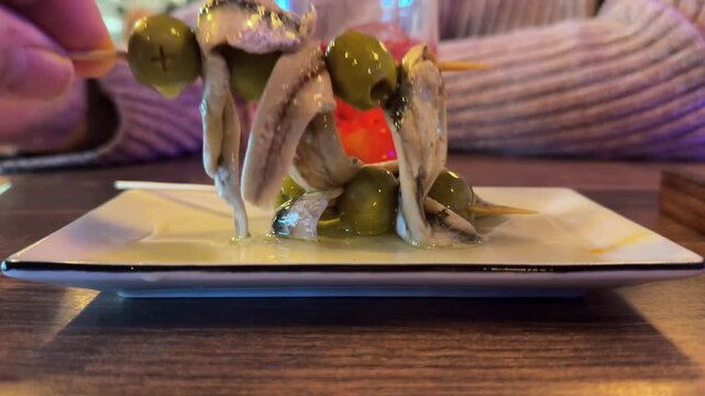 extreme closeup view of the hand of a man picking up a skewer of green olives and anchoives inside a traditional spanish bar.