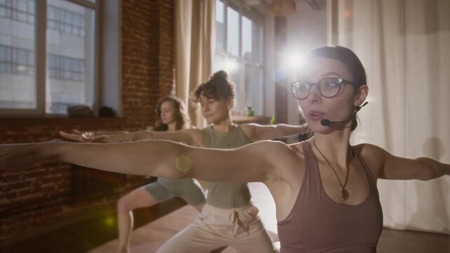 Medium close-up with lens flare of young Caucasian female yoga teacher with microphone instructing participants and demonstrating technique of warrior and triangle asanas, in sunlit red brick studio