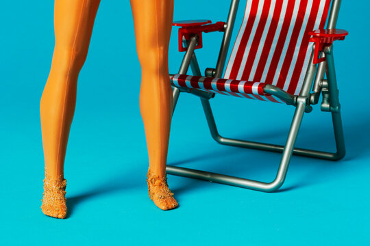 Sand covered doll feet and striped deck chair on blue background