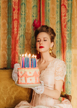 Woman in rococo dress holding birthday cake with lit candles indoors