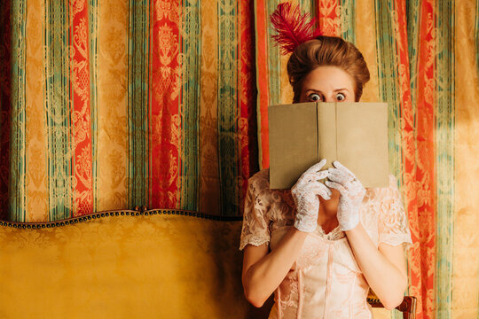 Woman in vintage Rococo dress reading a book and hiding face indoors