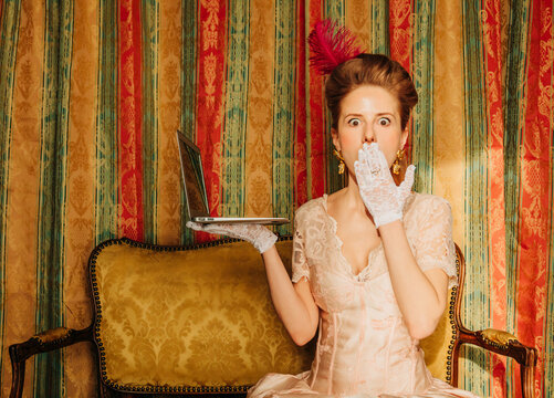 Surprised woman in rococo dress using laptop on vintage sofa indoors