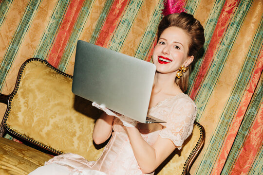 Smiling woman in vintage rococo dress using laptop on ornate sofa