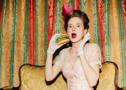 Surprised woman in vintage Rococo dress eating burger on ornate sofa