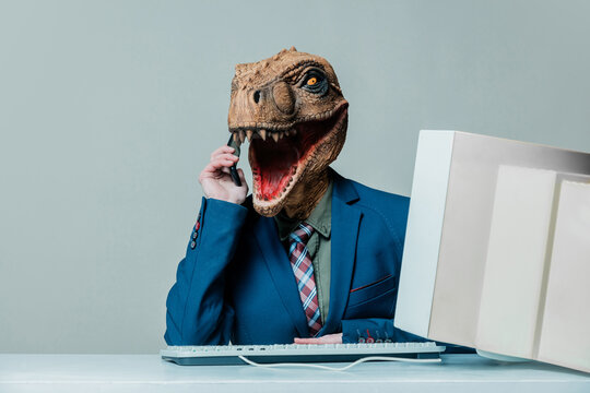Businessperson in dinosaur mask talking on phone at desk with old computer