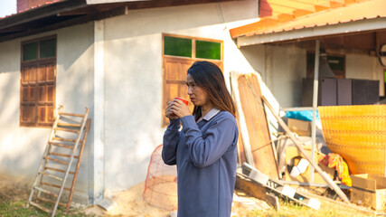 A woman stands outside a house holding a cup. She appears to be thoughtful while surrounded by building materials. The sun shines down on her and the surrounding area © Happy Photo