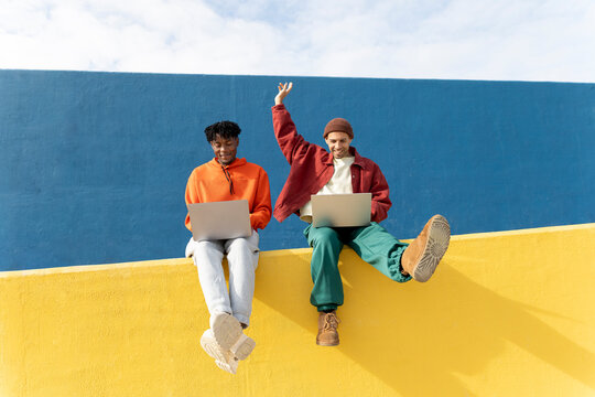 Friends sitting on colorful wall in Barcelona using laptops celebrating success