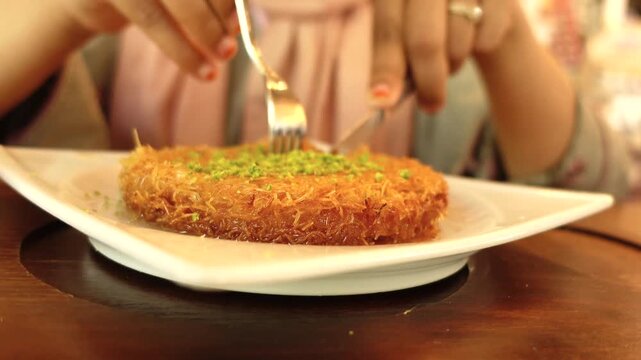 A captivating close-up of a person skillfully cutting and savoring a delicious kunefe with a fork and knife, golden brown dessert, covered with crushed pistachios, a traditional Middle Eastern delight
