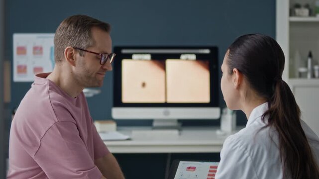 Female dermatologist using digital tablet while discussing skin mole with male patient during appointment in medical office, side angle shot with computer monitor display in background