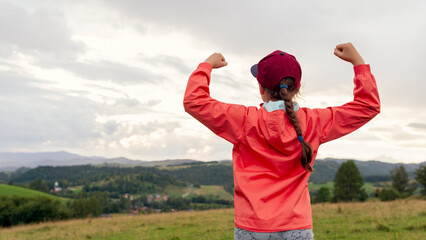Young girl hiker in orange jacket celebrating with raised arms while looking over scenic green valley and hills under dramatic cloudy sky