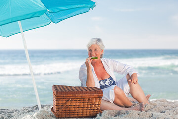 Senior woman sitting on shore biting cucumber wearing swimsuit beside teal umbrella, wicker basket