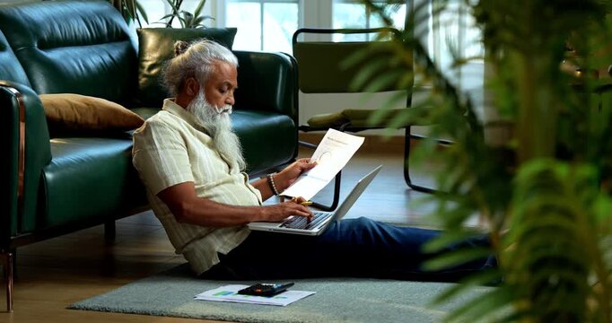 Indian mid age bearded man using laptop, calculator and reports for accounting sitting on carpet in modern home, stylish mature male managing finance, budget and financial statements indoors