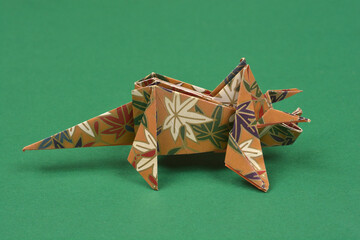 A single handmade paper origami triceratops with a brown and blue floral pattern stands on a solid dark green surface in a studio shot © nahhan