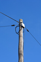Obraz premium A weathered wooden utility pole with various electrical wires and a white junction box stands against a clear and bright blue sky