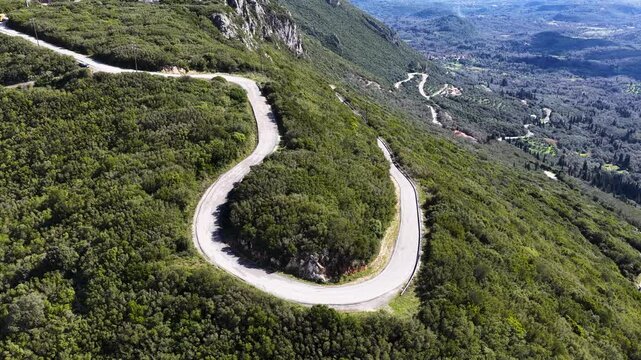 Drone flight over an empty asphalt mountain road with sharp hairpin curves surrounded by lush green trees on Corfu Island, Greece.