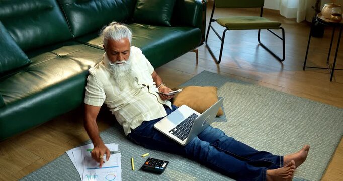 Indian mid age bearded man counting cash Indian rupees and relaxing between work sitting on carpet in modern home, stylish mature male managing household finance and budget indoors