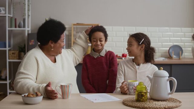 Medium shot of African American grandmother and granddaughter having conversation, drinking coffee and tea while little boy showing his grades