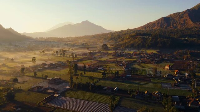 A stunning sunrise over Mount Batur and Mount Agung in Bali reveals mist-covered settlements and lush green plantations. The early light casts a serene glow over the breathtaking landscape.