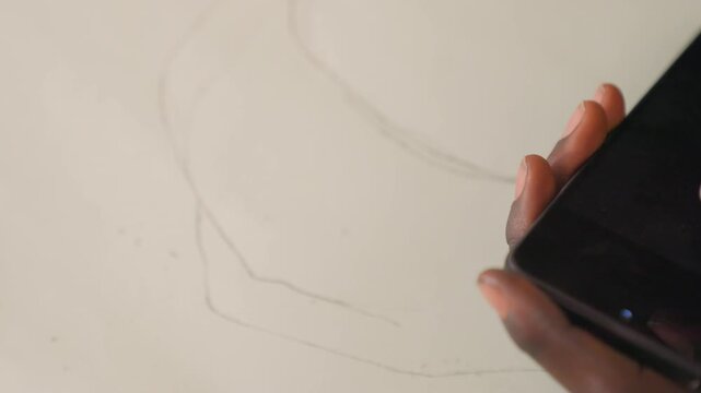 Black hand holding phone reference sketch, closeup of darkskinned hand viewing highcontrast image on screen while faint pencil outlines trace portrait on white paper, quiet focused studio mood,