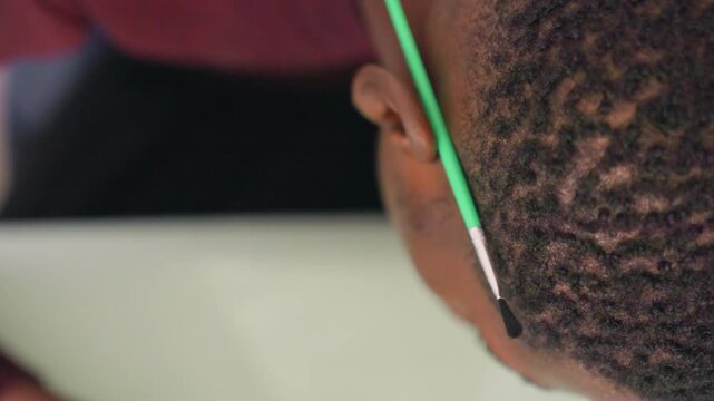 Closeup black hair painting with brush, quiet classroom scene of young student seated at table, gentle brush tip tracing scalp and hairline, textured waves and curls captured in soft light, attentive