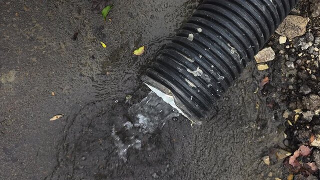 Water gushing from a black corrugated drain pipe during a heavy downpour, creating a puddle on the asphalt with rocks and leaves scattered around the driveway