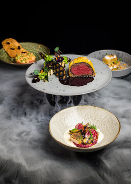 A mix of gourmet fusion fine dining dishes, including beef wellington, steak tartare, scallops and a mango cake dessert with dry ice creating a mist or smoke like effect