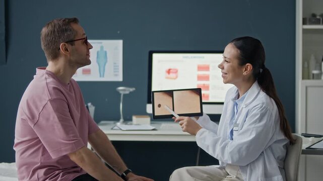Side angle medium shot of professional dermatologist pointing at skin mole photo on tablet screen while patient listening in clinic office with desktop monitor in background