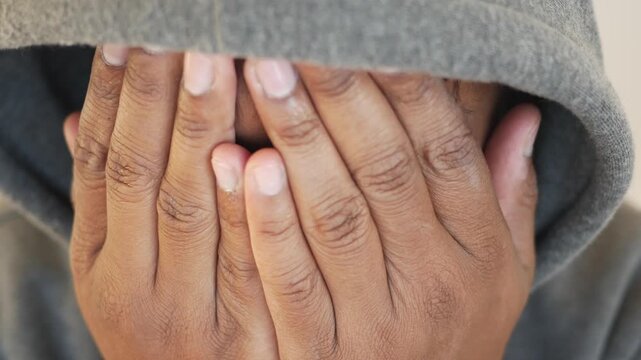 Close up video of young man in grey hoodie covering his face with hands and looking sad, portraying feelings of worry, stress, and anxiety
