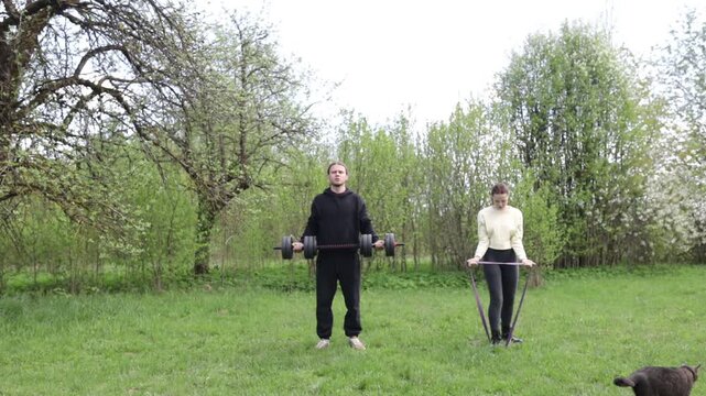 A young couple exercising outdoors in the summer, using a barbell and elastic band