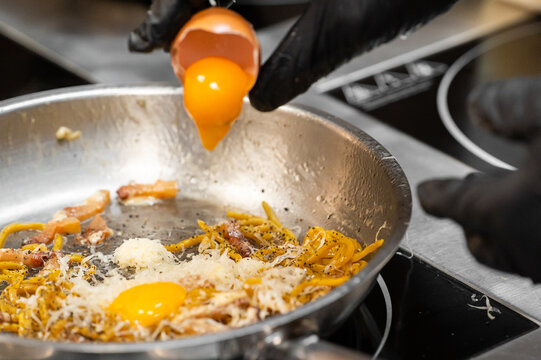 A chef in black gloves adds a fresh egg yolk to a stainless steel pan of creamy carbonara pasta with crispy pancetta and grated cheese, illustrating a professional cooking process
