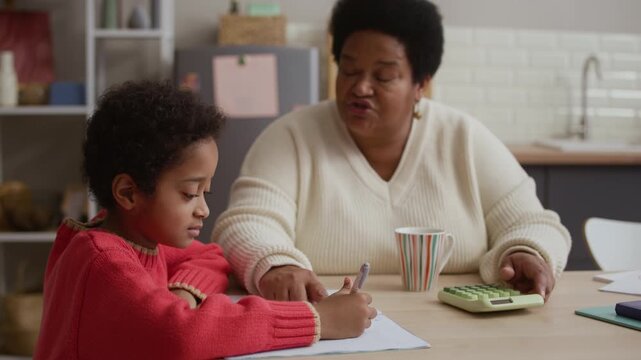 Little African American boy sitting at dining table and solving math problem while his grandmother helping him with calculation