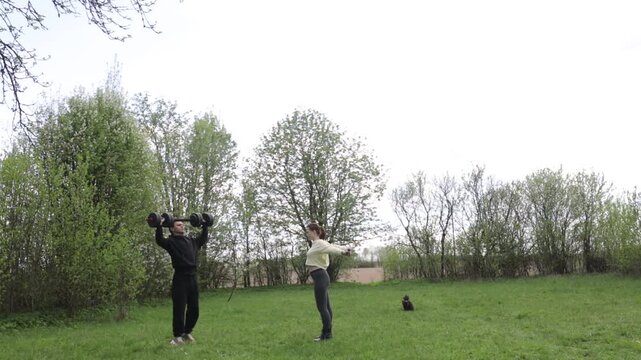 A young couple exercising outdoors in the summer, using a barbell and elastic band