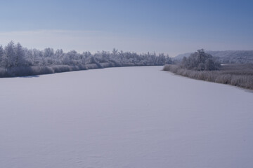 Icy river surface surrounded by dry reeds and frost covered forest. Minimal cold season landscape.