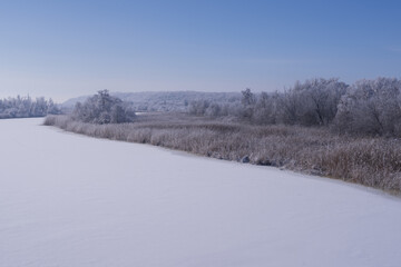 Icy river surface surrounded by dry reeds and frost covered forest. Minimal cold season landscape.