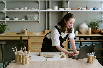 Young adult Caucasian woman standing at pottery studio table using laptop, surrounded by ceramic tools and clay, engaging in creative process with pottery pieces visible on shelves © AnnaStills