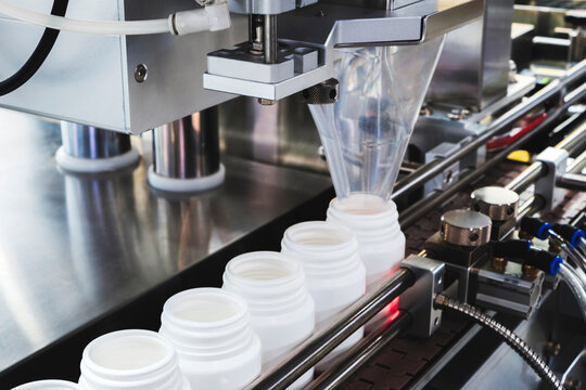 filling and packaging machine at pharmaceutical factory fills white plastic jars with tablets or ampoules containing dietary supplements. containers are lined up on conveyor belt.
