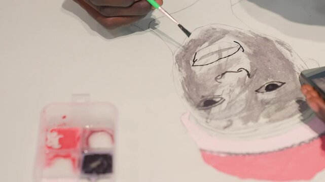 Closeup black artist adding finishing strokes to lips, green brush shaping small mouth detail on monochrome portrait, pink hat accent and paint case nearby, warm studio mood with careful playful touch