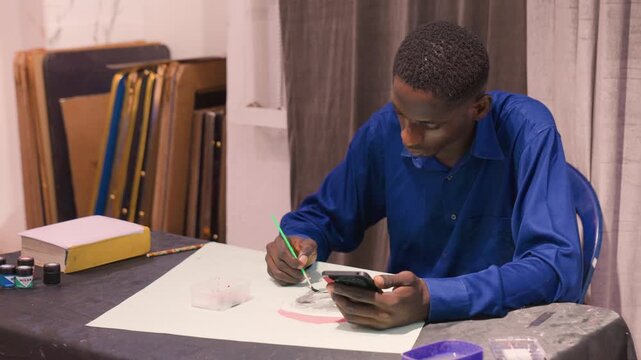 Black artist sketching portrait using phone reference, seated at studio table, blue shirt, white paper on blackcovered table, frames stacked in background, jars of ink, careful pencil and small brush