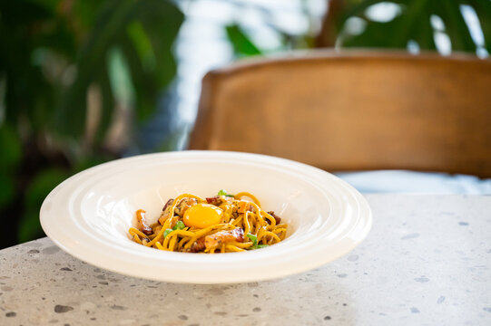 A bowl of spaghetti carbonara topped with a fresh egg yolk, served on a marble table in a bright, modern restaurant setting with lush green plants in the background.