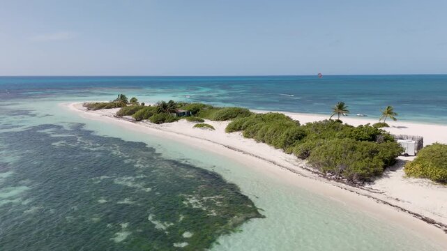 Aerial drone shot of a pristine tropical sandbar with palm trees, white sand, and crystal clear aqua water, kite surfer and boats visible near the shoreline under a clear blue sky